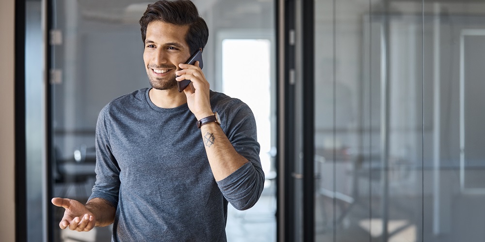 En mand i en grå og blå skjorte står i et rum, der er malet i næsten den samme farve. Han taler i telefon. Det ser ud til, at han modtager gode nyheder, da han smiler glad.