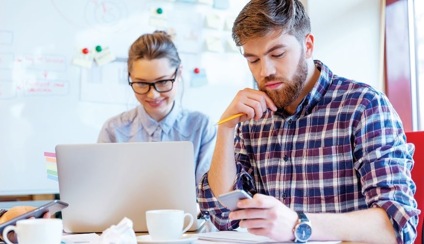 A young man and a young woman are sitting in a colorful office. They are both working on a project. The man is focussing on paperwork while the woman works happily on her laptop.